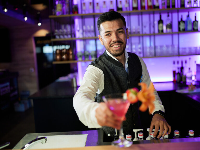 Barkeeper stellt Cocktail auf die Theke der Bar im 4-Sterne-Wellnesshotel Hüttenhof Barkeeper stellt Cocktail mit Blume auf Theke der modernen, violett beleuchteten Bar
